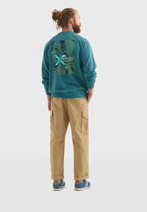 Sudadera azul verdosa con un estampado gráfico en la parte posterior, combinada con pantalones cargo beige que tienen bolsillos laterales y zapatillas azules con detalles en marrón claro.