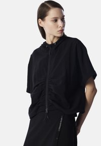 Veste noire légère cintrée avec capuche, dotée d'une fermeture éclair sur le devant, de détails froncés et de manches courtes.