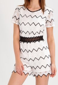 Vit spetsromper med svarta zigzag-mönster och scalloped kanter. Korta ärmar och en midja i svart spets. Tjock och semi-transparent design.