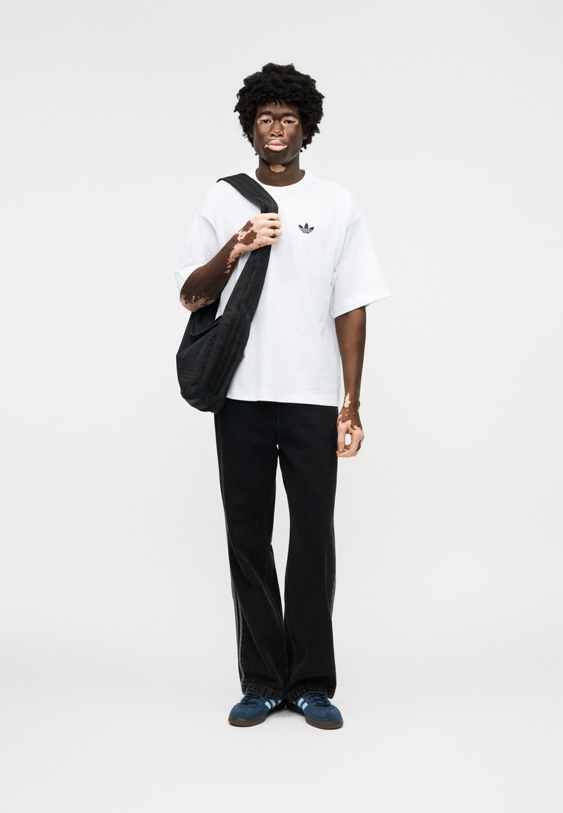 Junge Person mit Vitiligo, trägt weißes Adidas-T-Shirt, schwarze Hose, blaue Sneakers und eine schwarze Umhängetasche, steht vor einem schlichten weißen Hintergrund.