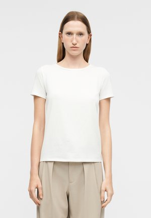 Kobieta z prostymi jasnobrązowymi włosami ubrana w prostą białą koszulkę z krótkim rękawem i beżowe plisowane spodnie, stoi na tle jasnoszarego tła.