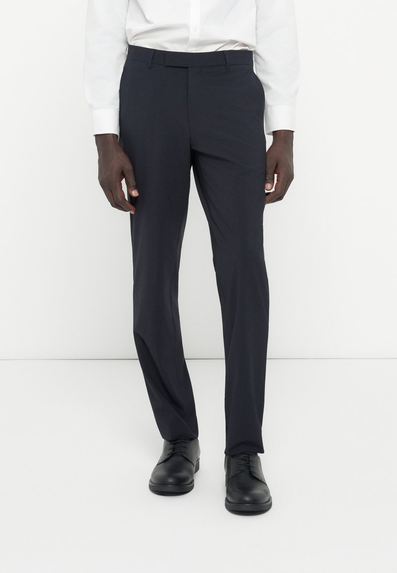 Marineblaue Anzughose aus glattem Stoff, mit taillierter Passform und flachem Vorderteil. Kombiniert mit schwarzen Lederschuhen für einen formellen Look.