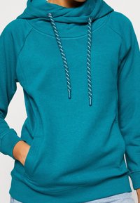 Sweat à capuche couleur sarcelle avec une coupe décontractée, doté de cordons à rayures blanches, d'une poche frontale et de poignets côtelés. Matière douce et texturée.