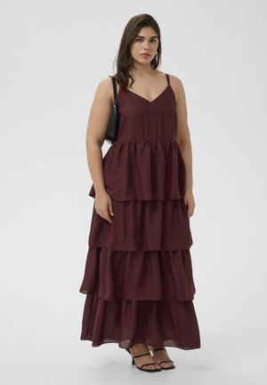 Naine, kes kannab burgundia tooni kihilist maksikleid koos õhukeste rihmadega, musta õlakotti ja musti rihmikujulisi kontsakingi, seisab ühevärvilise tausta ees.