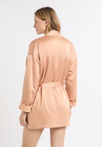 Satynowy szlafrok kimono w miękkim odcieniu brzoskwini, z długimi rękawami i paskiem do wiązania; posiada miękkie mankiety i sięga do kolan.