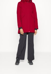 Parka impermeable roja con capucha, dos bolsillos frontales y detalles de cremallera. Combinado con pantalones grises de corte relajado y zapatillas deportivas negras.