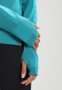 Tee in Türkis mit langen Ärmeln, Daumenlöchern, strukturierten Einsätzen und taillierter Passform, kombiniert mit schwarzen Leggings. Glatte Stoffoberfläche.