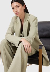 Completo su misura verde chiaro con colletto a bavero, pantaloni a gamba larga e dettagli con bottoni argentati. Indossato con un accessorio a più anelli. Sfondo di una sedia nera.