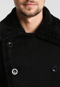 El abrigo negro tiene un cuello texturizado y suave, con dos botones negros y un botón con logo plateado cerca del pecho.