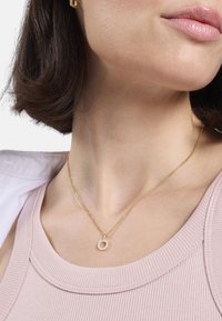 Collar de cadena de oro con un colgante circular que presenta pequeñas piedras claras, usado sobre una camiseta de rayas de color rosa claro. Textura suave, diseño sencillo.