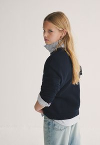 Maglione navy lavorato a maglia sovrapposto a una camicia azzurra a righe, con collo alto, vestibilità rilassata e jeans a lavaggio medio. Capelli lunghi e lisci.
