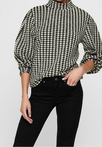 Pull noir et blanc à motif pied-de-poule avec col montant et manches bouffantes, porté avec un jean skinny noir, présentant une texture de tissu lisse.