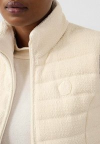 Gilet senza maniche color crema con tessuto a trama, chiusura con zip e dettaglio del logo sulla parte anteriore. Lo strato interno è una maglia leggera e liscia.