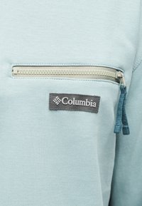 Ανοιχτό μπλε ύφασμα με οριζόντια τσέπη με μπεζ φερμουάρ και γκρι ετικέτα με το λογότυπο Columbia κάτω από το φερμουάρ στο ένδυμα.