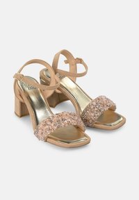 Sandalias de tacón beige con un tacón ancho; cuentan con una banda brillante y texturizada hecha de adornos claros y rosas en la plantilla.