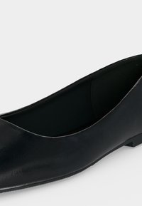 Chaussure ballerine plate pour femme en cuir noir avec petit talon, bout arrondi et finition lisse sur fond gris clair.