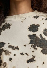 Sweatshirt beige à taches abstraites noires, avec un col rond côtelé et un tissu texturé, présenté de la clavicule vers le haut.