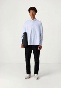 Νεαρός άνδρας στέκεται, φορώντας ανοιχτό μπλε πουκάμισο με κουμπιά, μαύρο παντελόνι, λευκά αθλητικά παπούτσια, κρατώντας μια διπλωμένη μαύρη ζακέτα στο αριστερό του χέρι.