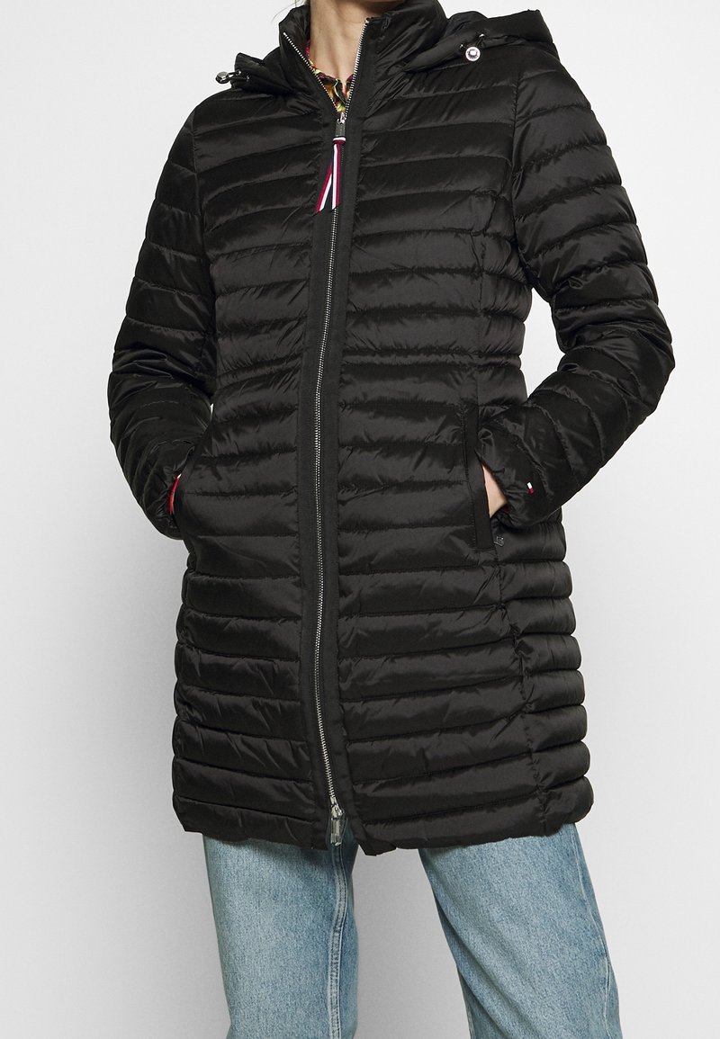 Doudoune noire avec motif matelassé horizontal, fermeture éclair complète, poches zippées et capuche amovible. Le matériau semble lisse et brillant.