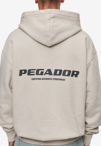 Bež dukserica s velikim crnim logom "PEGADOR" na leđima, uz tekst "CERTIFIED AUTHENTIC STREETWEAR" ispod u manjem fontu.