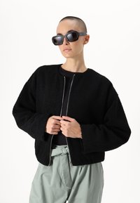 Giacca nera, texture, con collo rotondo e chiusura con zip, abbinata a pantaloni verde a vita alta e occhiali da sole neri oversize.