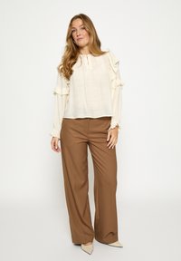 Kreemjas volangidega pluus kolmeveerandvarrukatega, kombineeritud kõrge vöökohaga pruunide laiade säärtega pükstega ning beežide teravate ninaosadega kontsakingadega.