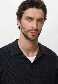 Camiseta polo acanalada en un azul marino oscuro con cuello en V y mangas cortas, que cuenta con una tela texturizada y un cuello doblado. Camiseta blanca debajo.