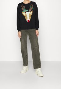 Felpa nera con un grafico di renna con corna, agrifoglio e un naso rosso, abbinata a jeans larghi verde oliva e sneakers bianche.