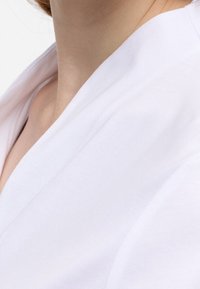 Abbigliamento in cotone bianco con design a scollo a V, caratterizzato da una texture liscia e cuciture minime, con un primo piano sulla zona del collo.