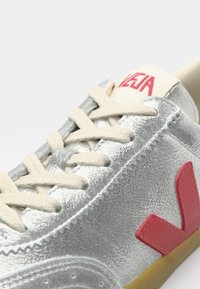 Silver leather sneaker with beige laces, featuring a pink V logo on the side and a textured detail across the upper. Rubber sole.