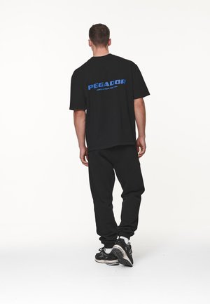 Camiseta negra de gran tamaño con el logotipo azul "PEGADOR" en la espalda, combinada con pantalones deportivos negros y zapatillas negras. Diseño simple y casual.
