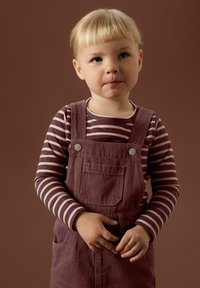 Jeune enfant aux cheveux blonds portant un t-shirt à rayures à manches longues et une salopette bordeaux, se tenant les mains jointes devant un fond marron.