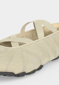 Μπεζ παπούτσι slip-on με σταυρωτούς ελαστικούς ιμάντες, ραμμένο ανώτατο μέρος από σουέντ και μαύρη υφασμάτινη λάστιχη σόλα.