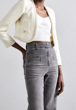 Frau trägt eine cremefarbene Cropped-Jacke mit Perlbesatz, ein weißes Cropped-Top und hoch taillierte graue Jeans mit vorderen aufgesetzten Taschen.