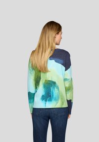 Frau mit langen blonden Haaren, die einen Pullover mit blau-grünem abstraktem Muster und eine dunkelblaue Jeans trägt, steht vor einem schlichten grauen Hintergrund.