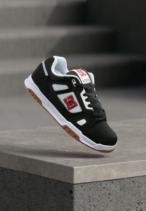 Sort-hvid DC skate sko med rødt logo, gummisål og sorte snørebånd, svævende over en betonforkant i blødt lys.