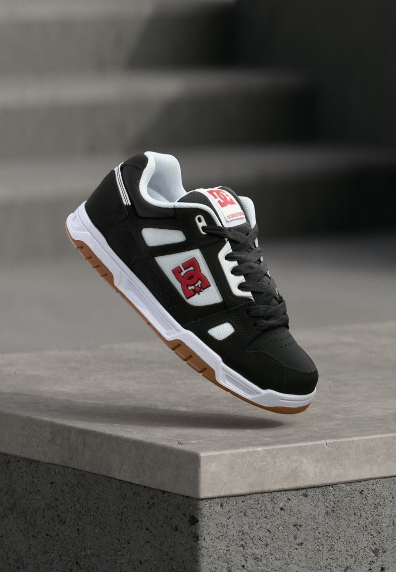 Ασπρόμαυρο παπούτσι DC για σκέιτ με κόκκινο λογότυπο, λαστιχένια σόλα και μαύρα κορδόνια, να αιωρείται πάνω από μια τσιμεντένια άκρη σε απαλό φως.
