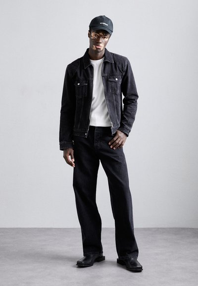 Veste en denim noir avec poches, t-shirt blanc, jeans noirs et chaussures plates noires. Une casquette noire avec texte complète la tenue.