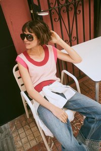 Femme aux cheveux courts bruns portant des lunettes de soleil, un top sans manches en maille rose et rouge, un jean bleu et une chemise blanche nouée à la taille, assise à une table.