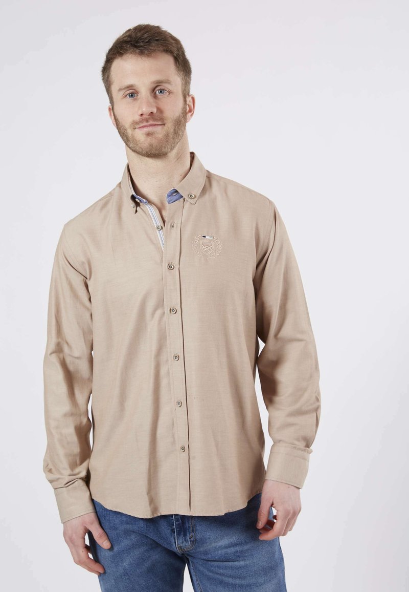 Chemise beige à manches longues avec un col boutonné, rayures subtiles sur la patte de boutonnage intérieure, logo brodé sur la poitrine et texture de tissu lisse.