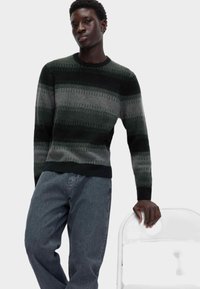 Strickpullover in dunkelgrün und grau mit horizontalen Streifen, mit Rundhalsausschnitt, kombiniert mit dunklen Jeans. Das Modell lehnt sich an einen weißen Stuhl.