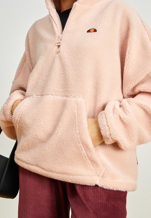 Person trägt einen hellrosa Fleece-Pullover mit halbem Reißverschluss, Kängurutasche und kleinem gesticktem Logo, kombiniert mit bordeauxfarbenen Hosen.