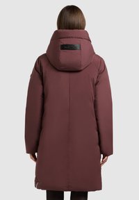 Pikk tumebordoopunane puffer jope suure reguleeritava kapuutsi, kaubamärgiga tagatasku ja allääres ventilatsiooni jaoks tõmblukkudega.