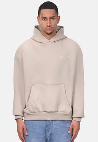Beige hooded sweatshirt with front pocket, ribbed cuffs, and hem. Features small white embroidered logo on the chest. Soft fabric texture.