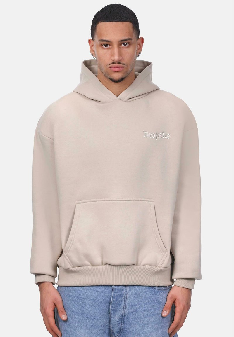 Beige hættebluse med frontlomme, ribbede manchetter og kant. Har lille hvid broderet logo på brystet. Blød stoftekstur.