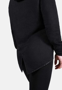 Fekete, hosszú ujjú felső, textúrált felülettel, oldalsó cipzárral és ívelt aljjal, fekete leggings-szel párosítva.