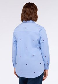 Camicetta azzurra chiara in cotone con colletto a punta, caratterizzata da piccole ricami a forma di cuore rossi sparsi e polsini con bottoni.