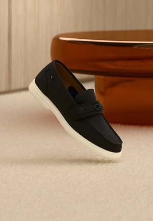 Μαύρο σοσόνι από σουέντ με λευκή σόλα που αιωρείται πάνω από μπεζ χαλί, καφέ στρογγυλό τραπέζι σε φόντο απαλού φωτισμού.