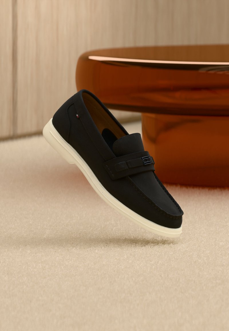 Μαύρο σοσόνι από σουέντ με λευκή σόλα που αιωρείται πάνω από μπεζ χαλί, καφέ στρογγυλό τραπέζι σε φόντο απαλού φωτισμού.