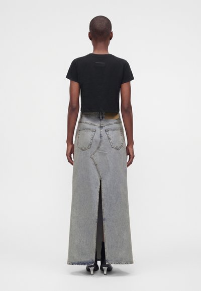 Άτομο που φοράει μαύρο cropped t-shirt, μακριά ανοιχτόγρια τζιν φούστα με σχίσιμο στο πίσω μέρος και μαύρα παπούτσια με τακούνι, στέκεται με την πλάτη σε λευκό φόντο.